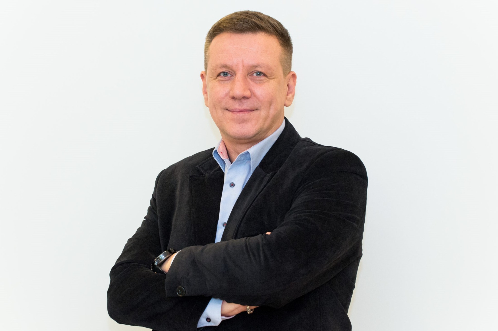 Portret mężczyzny w czarnej marynarce i błękitnej koszuli, z założonymi rękami na tle białej ściany.