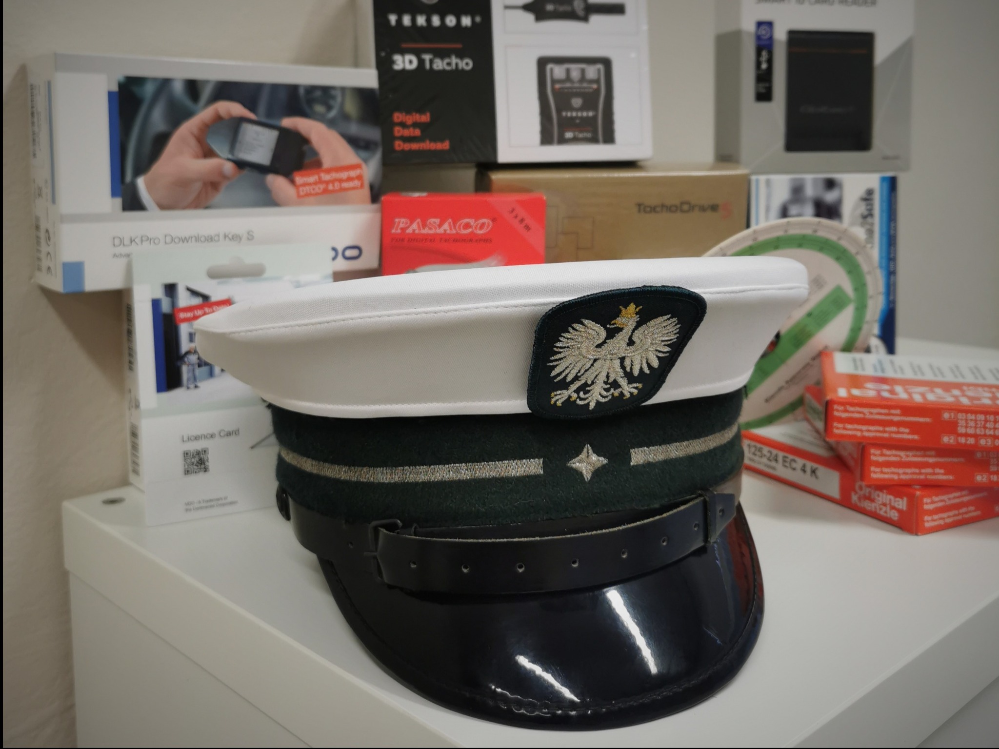 Czapka służbowa z orłem na tle pudełek z tachografami i akcesoriami do nich.