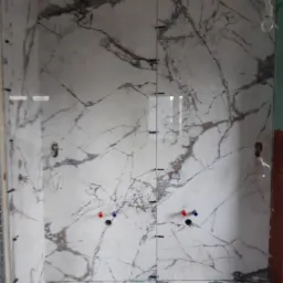 Ściana łazienki z białych płytek imitujących marmur, widoczne otwory na armaturę i narzędzia glazurnicze na podłodze.