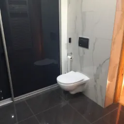 Nowoczesna toaleta podwieszana z czarnym panelem spłukującym na ścianie wyłożonej białymi płytkami imitującymi marmur, obok czarnej szklanej kabiny prysznicowej i ciemnej, błyszczącej podłogi.