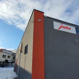 Zewnętrzne kamery monitoringu zainstalowane na rogu szarego budynku z pomarańczowym akcentem, widok z dołu na tle pochmurnego nieba.