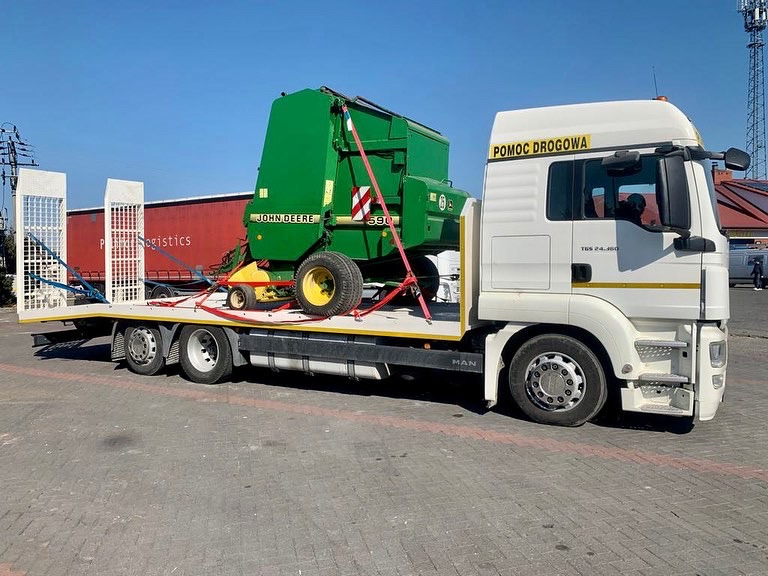 Biała ciężarówka z napisem 'POMOC DROGOWA' przewozi na niskopodwoziowej platformie zieloną prasę rolniczą John Deere 590 zabezpieczoną czerwonymi pasami.