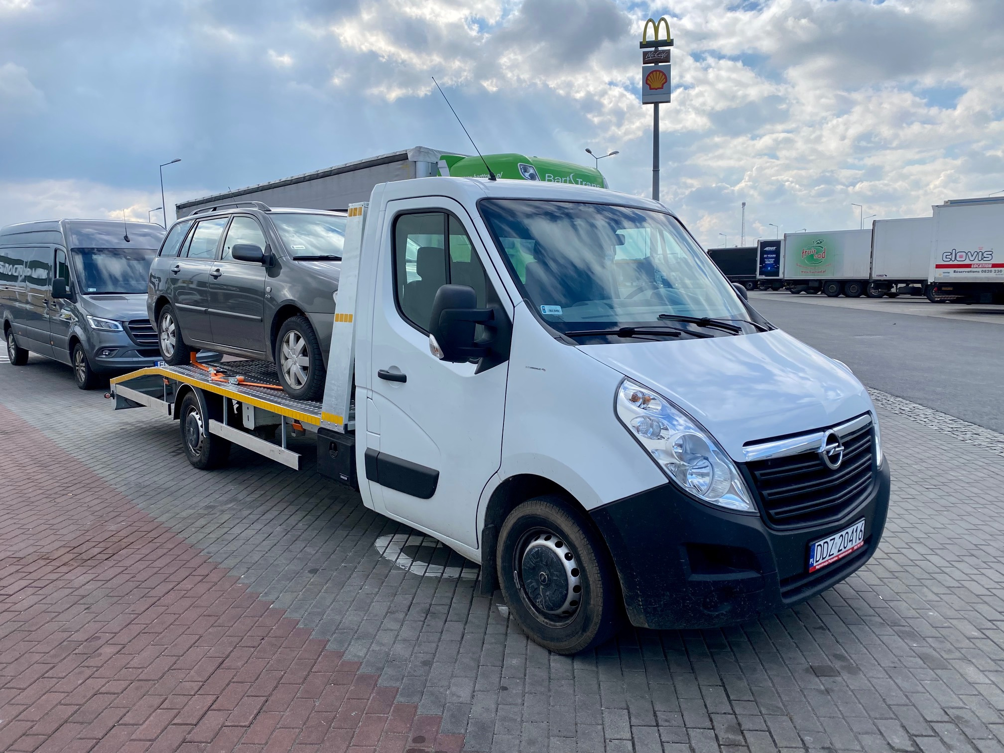 Biała laweta z szarym kombi na platformie, zaparkowana na brukowanym placu postojowym z widocznymi tirami w tle i znakiem McDonald's oraz Shell.