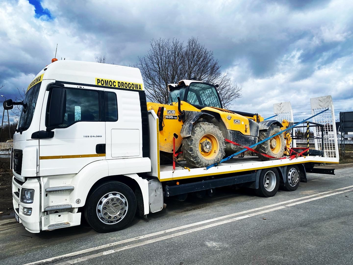 Biała laweta z napisem 'Pomoc Drogowa' przewozi żółtą maszynę budowlaną JCB 535-95 zabezpieczoną pasami transportowymi na tle pochmurnego nieba.