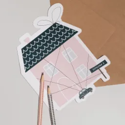 Ręcznie robiona kartka w kształcie różowego domu z czarnym dachem, stylizowana na zaproszenie, z narysowanym samochodem, adresem 'Tofolowa 8' i dwoma ołówkami na jasnym tle.