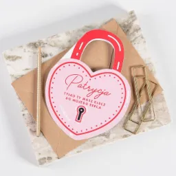 Kartka walentynkowa w kształcie kłódki z napisem 'Patrycja, tylko ty masz klucz do mojego serca' leżąca na kopercie obok złotego ołówka i spinaczy na marmurowej płytce.