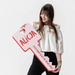 Uśmiechnięta młoda kobieta trzyma w rękach duży, różowy, dekoracyjny klucz z napisem 'Alicja' i sentencją 'Przysłałam klucz do mego serca', na białym tle.