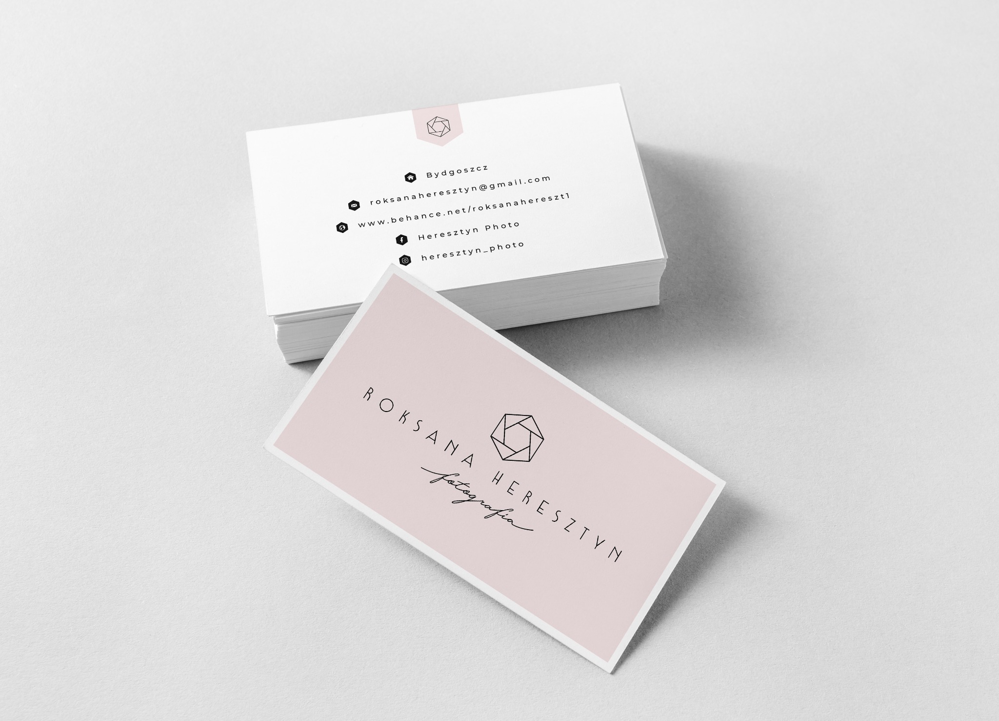 Stos wizytówek z minimalistycznym logo i danymi kontaktowymi na jasnoróżowym tle, ułożony na białym tle.