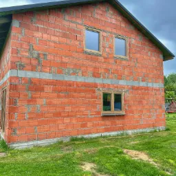 Surowy budynek z czerwonej cegły z zamontowanymi oknami z ramami w kolorze drewna, widoczne szare nadproża i fragment dachu, na pierwszym planie trawnik.