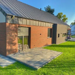 Nowoczesny dom jednorodzinny z ceglaną elewacją, szarą blachodachówką i częściowo zasłoniętymi roletami zewnętrznymi, drabina oparta o ścianę, widoczny fragment tarasu z kostki brukowej i zadbany...