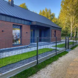 Nowoczesny dom jednorodzinny z cegły klinkierowej, grafitowym dachem i ogrodzeniem panelowym z podmurówką betonową, widok z perspektywy ścieżki.