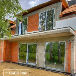 Surowy stan budynku w trakcie montażu stolarki okiennej; widoczne cegły, beton, częściowo zamontowane okna z szarymi ramami i odbijającym się otoczeniem, ziemia przed budynkiem.