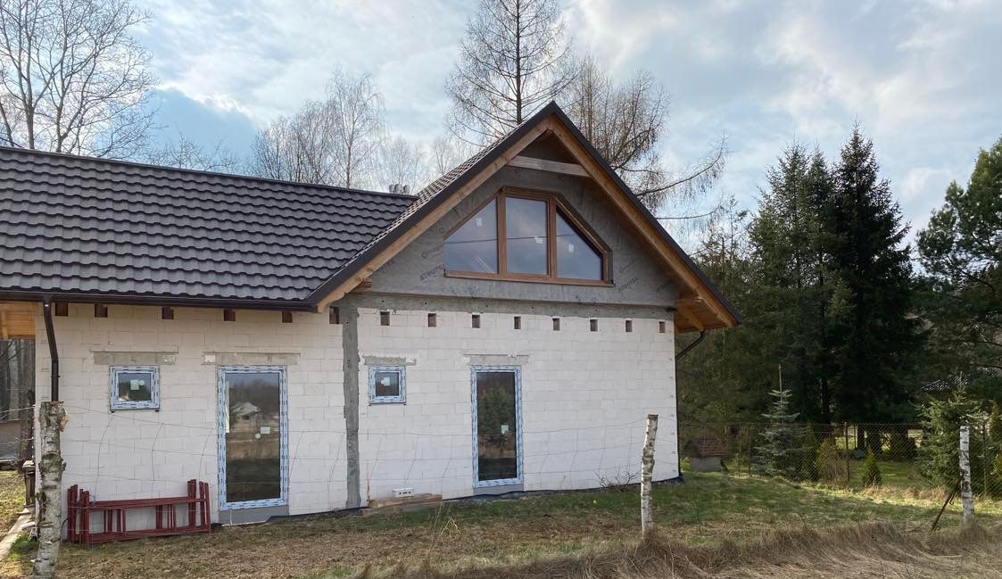 Dom w stanie surowym otwartym z zamontowanymi oknami w kolorze winchester i oknem trójkątnym na poddaszu, widoczna drabina i niezabezpieczone otwory po oknach.