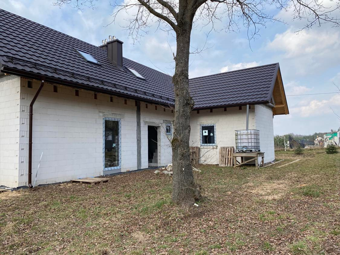 Dom w trakcie budowy z białego bloczka, z zamontowanymi oknami z niebieską folią ochronną, brązowy dach z blachodachówki, widoczne palety i zbiornik na wodę.