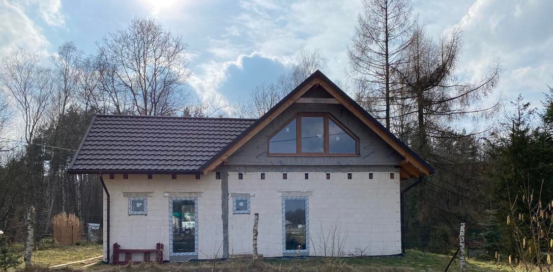 Dom w trakcie budowy z brązowym dachem, oknami w kolorze winchester i ścianami z białego bloczka, widoczne drzewa w tle.