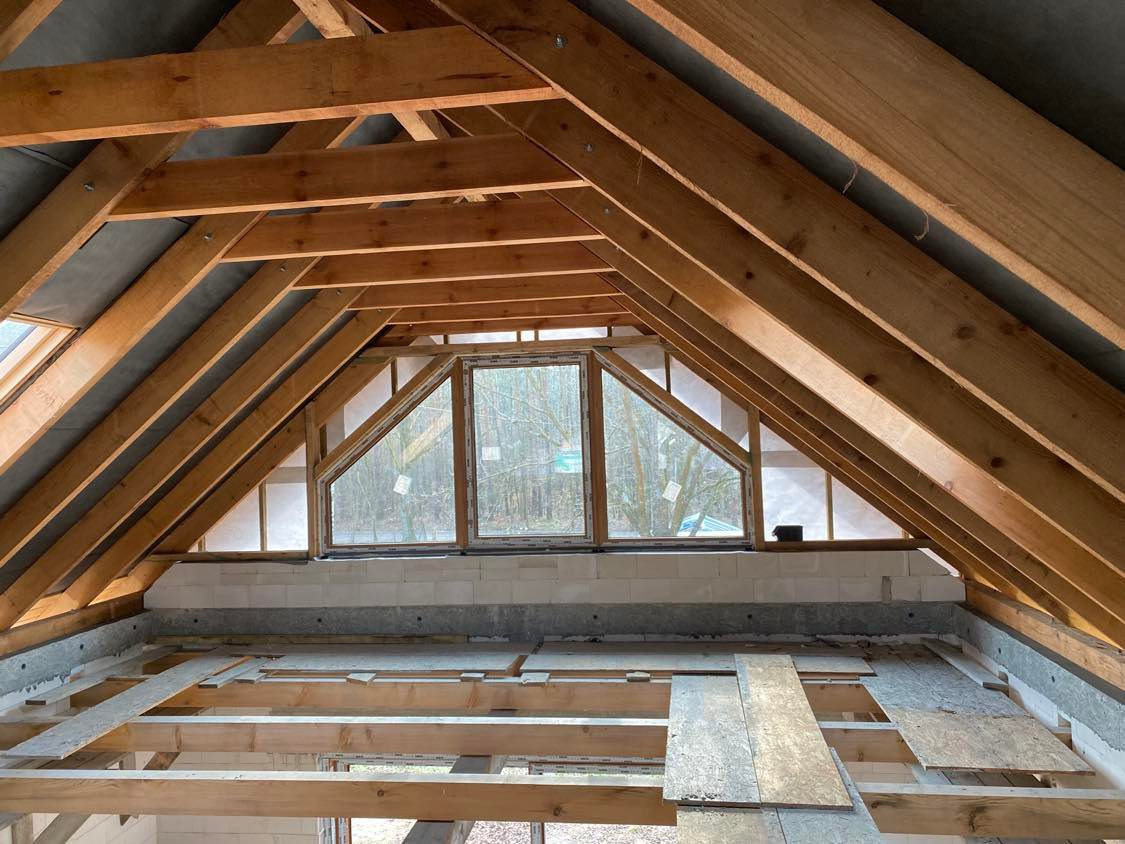 Trzy trapezoidalne okna w szczycie dachu, widok na drzewa, surowa konstrukcja drewniana dachu w trakcie budowy, bloczki betonowe poniżej okien.