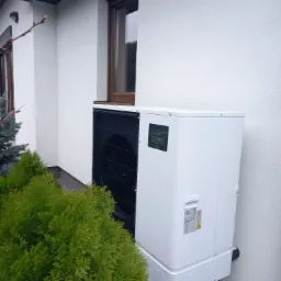 Biała pompa ciepła typu monoblok, zainstalowana na zewnątrz budynku z jasną elewacją, widoczna z boku, obok zielonych krzewów i okna z brązową ramą.