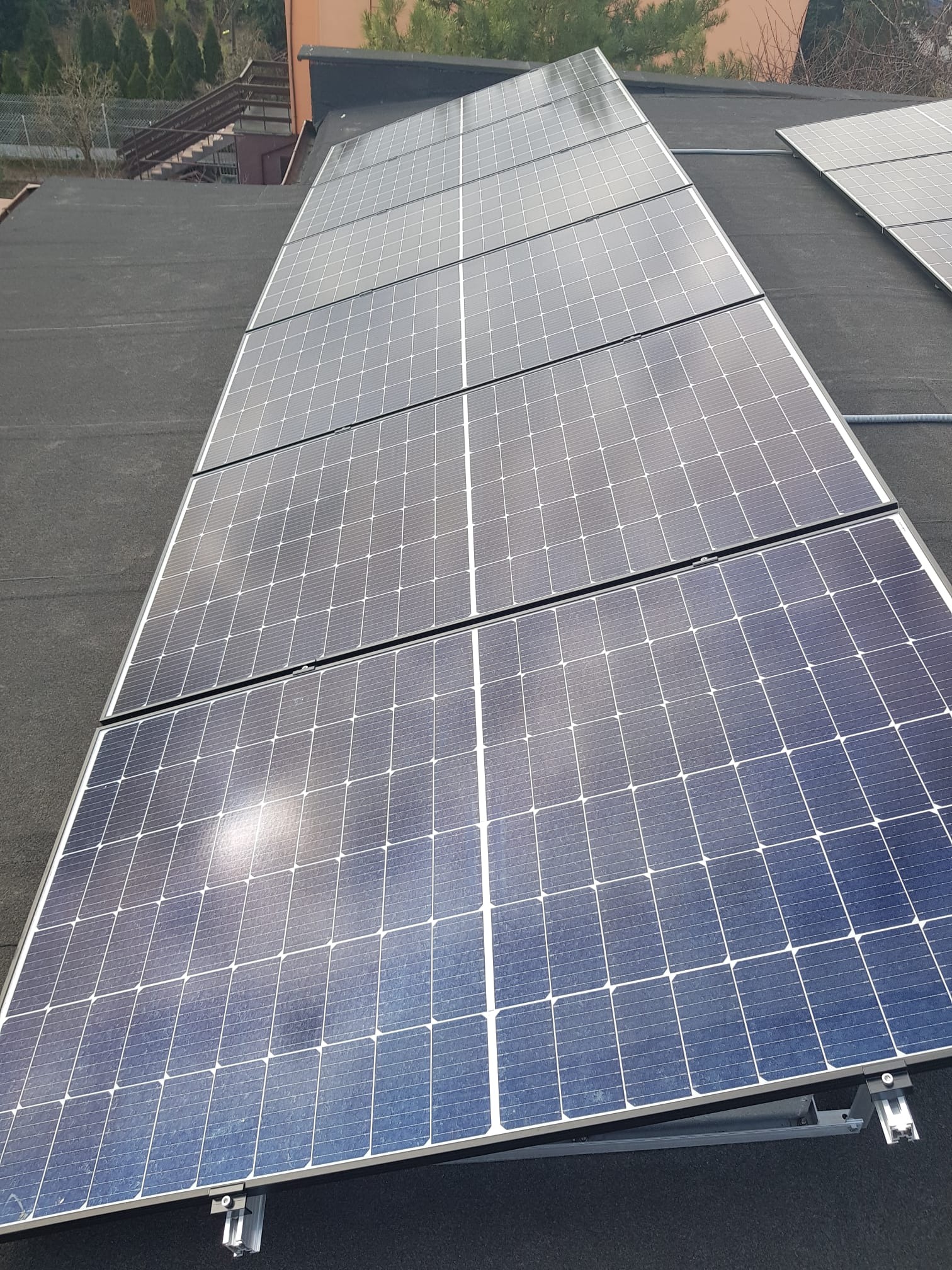 Panele fotowoltaiczne zamontowane na płaskim dachu pokrytym papą, widoczne elementy mocujące i odbicie nieba w panelach.