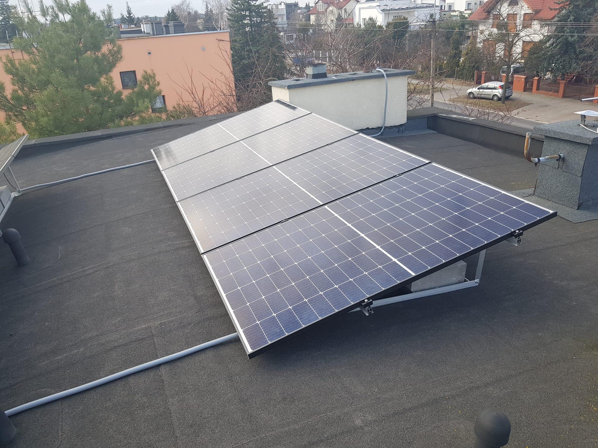 Cztery panele słoneczne zamontowane na płaskim, czarnym dachu z widocznymi elementami konstrukcyjnymi i rurą ochronną kabli, w tle budynki mieszkalne i drzewa.