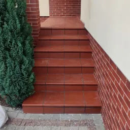 Wykończone płytkami schody wejściowe do domu z ceglaną elewacją i ozdobnym drzewkiem iglastym po lewej stronie.