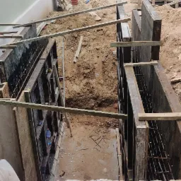 Przygotowanie szalunków i zbrojenia pod fundament, widoczne drewniane podpory, wykop ziemny oraz fragment ściany budynku w surowym stanie.