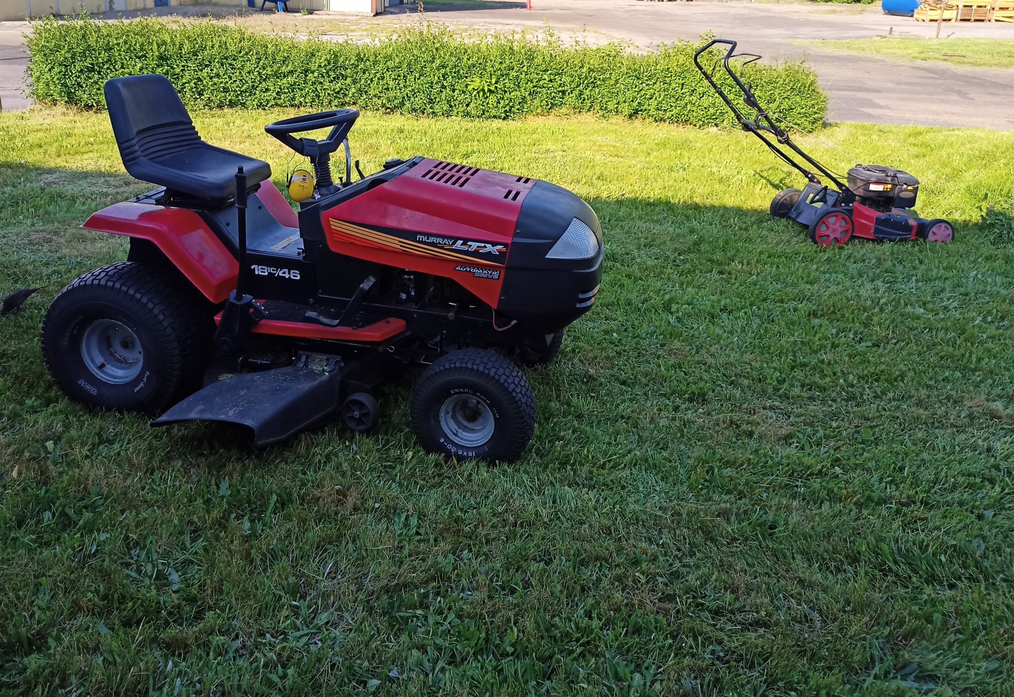 Czerwony traktorek do koszenia trawy Murray LTX i kosiarka spalinowa na trawniku przed krzewami.