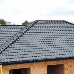 Ciemnoszary dach z dachówki ceramicznej na surowym domu z cegły, widok z góry na tle pochmurnego nieba i zieleni drzew.