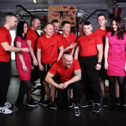 Grupa uśmiechniętych trenerów personalnych EgoBody we Wrocławiu, ubranych w czerwone koszulki z logo firmy, pozuje w siłowni na tle sprzętu do ćwiczeń.