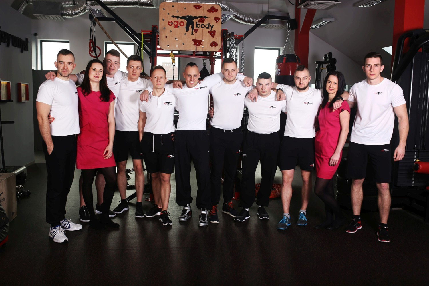 Grupa uśmiechniętych osób, w tym kobiety w czerwonych sukienkach, i mężczyźni w białych koszulkach i czarnych spodniach/szortach, pozujących w siłowni pod logo EgoBody