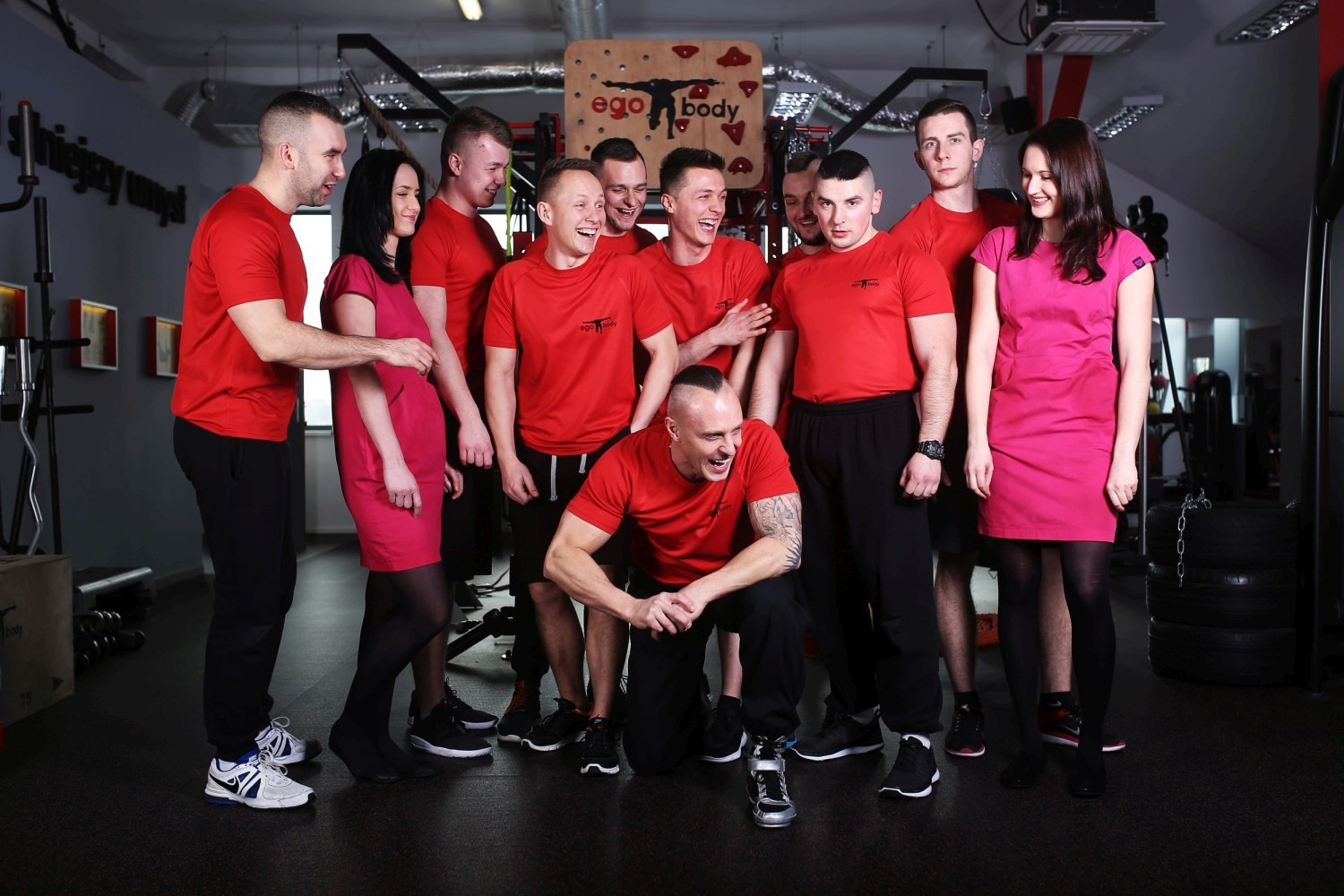 Grupa uśmiechniętych trenerów personalnych EgoBody we Wrocławiu, ubranych w czerwone koszulki z logo firmy, pozuje w siłowni na tle sprzętu do ćwiczeń.