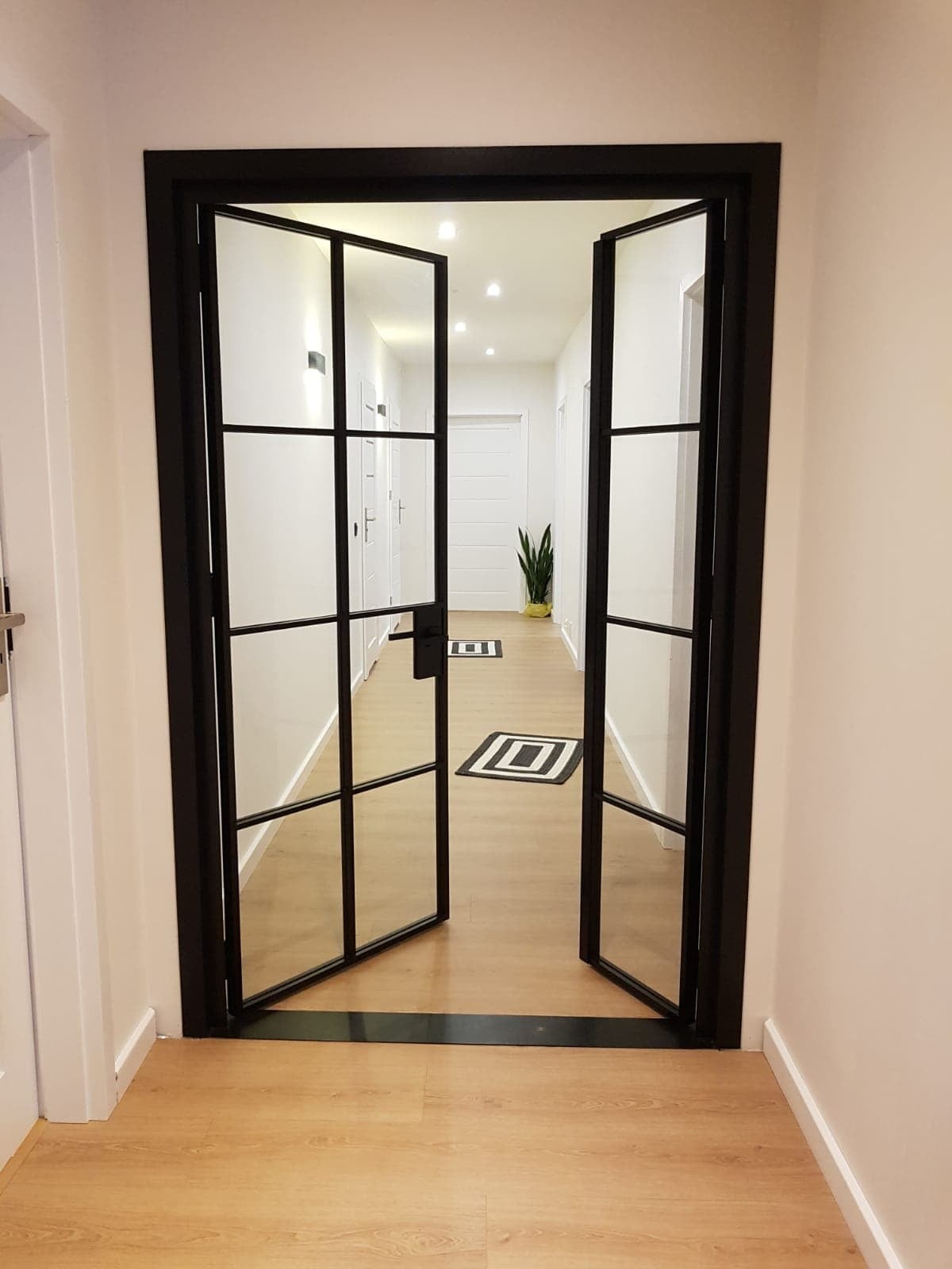 Dwuskrzydłowe drzwi wewnętrzne w stylu loft z czarnymi szprosami i przezroczystym szkłem, otwarte do korytarza z drewnianą podłogą i dwoma dywanikami w geometryczny wzór.