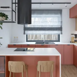 Nowoczesna kuchnia w odcieniach koralu z marmurowym blatem, drewnianymi krzesłami barowymi i minimalistycznym oświetleniem, okno z roletą rzymską z widokiem na las.