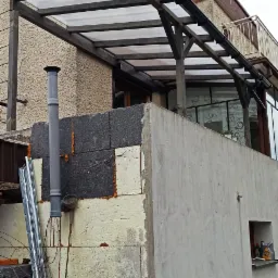 Widok z dołu na drewnianą konstrukcję z przezroczystym zadaszeniem nad tarasem, z widocznymi rynnami, kominem i fragmentem elewacji budynku w trakcie ocieplania.