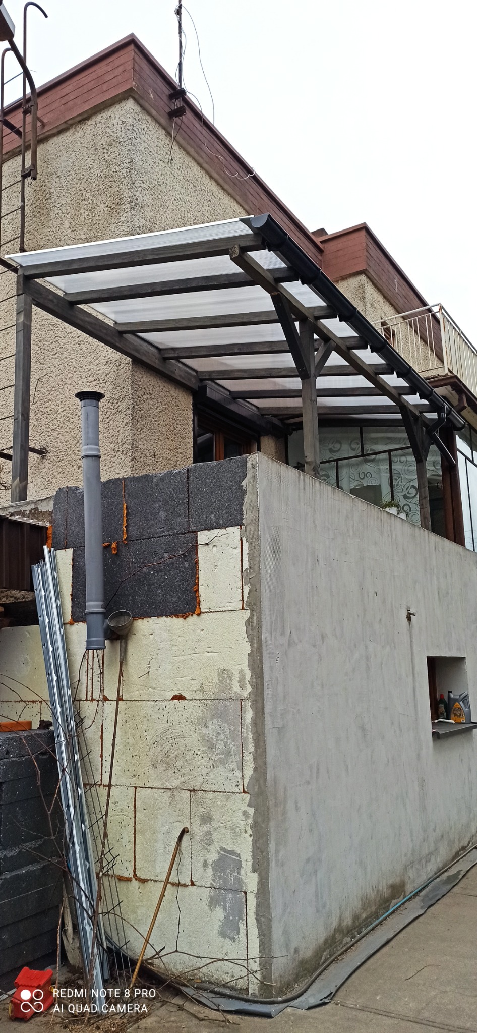 Widok z dołu na drewnianą konstrukcję z przezroczystym zadaszeniem nad tarasem, z widocznymi rynnami, kominem i fragmentem elewacji budynku w trakcie ocieplania.