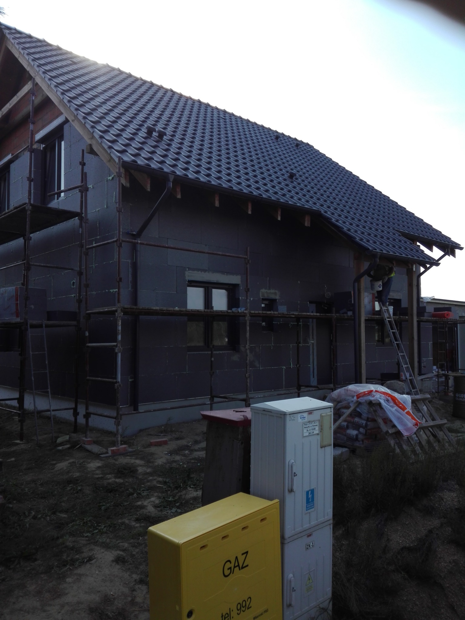 Dom w trakcie ocieplania i układania dachówki, widoczne rusztowania, materiały budowlane i skrzynki z oznaczeniami gazu i prądu na pierwszym planie.