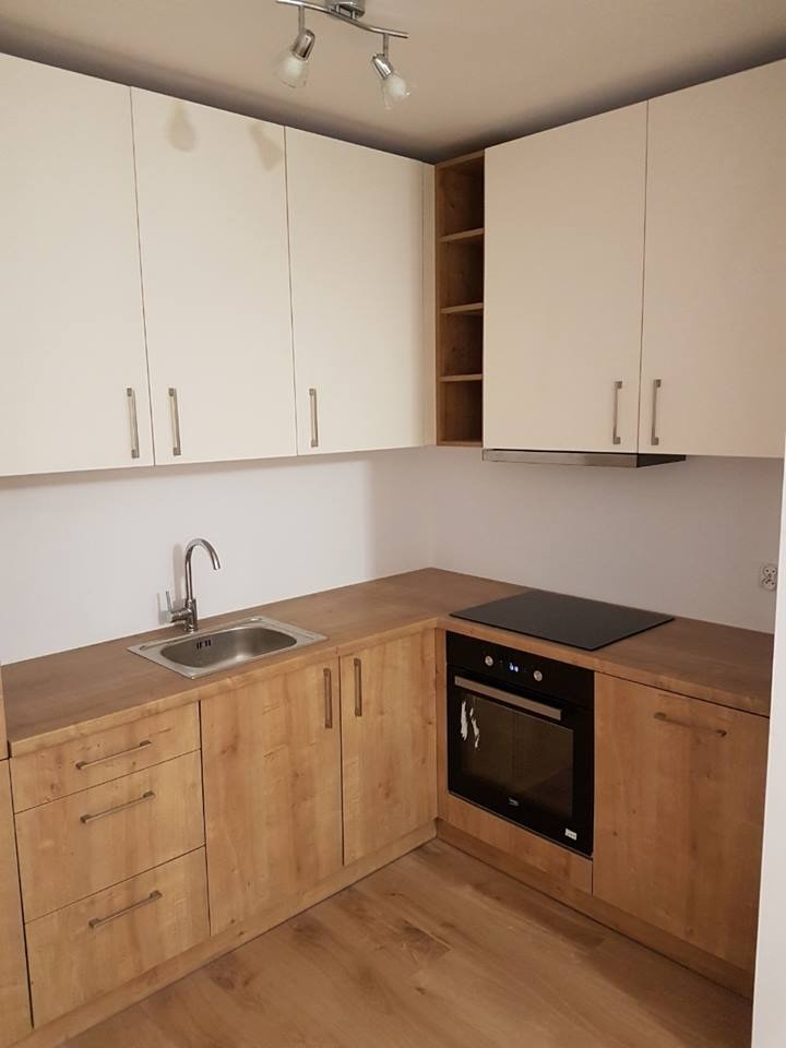 Jasna kuchnia z drewnianymi szafkami dolnymi i białymi górnymi, z wbudowanym piekarnikiem i płytą grzewczą, widok na zlew ze stali nierdzewnej i drewniany blat.