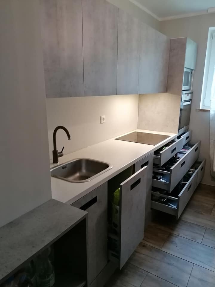 Szara kuchnia z blatem, otwartymi szufladami z wyposażeniem i widocznym zlewem z ciemną armaturą.