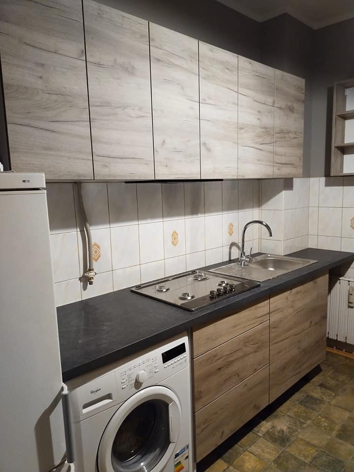 Kuchenna zabudowa z drewnianymi frontami, szarym blatem i płytą gazową, obok pralka automatyczna, widok na górne szafki w podobnym stylu, płytki ceramiczne na ścianie.