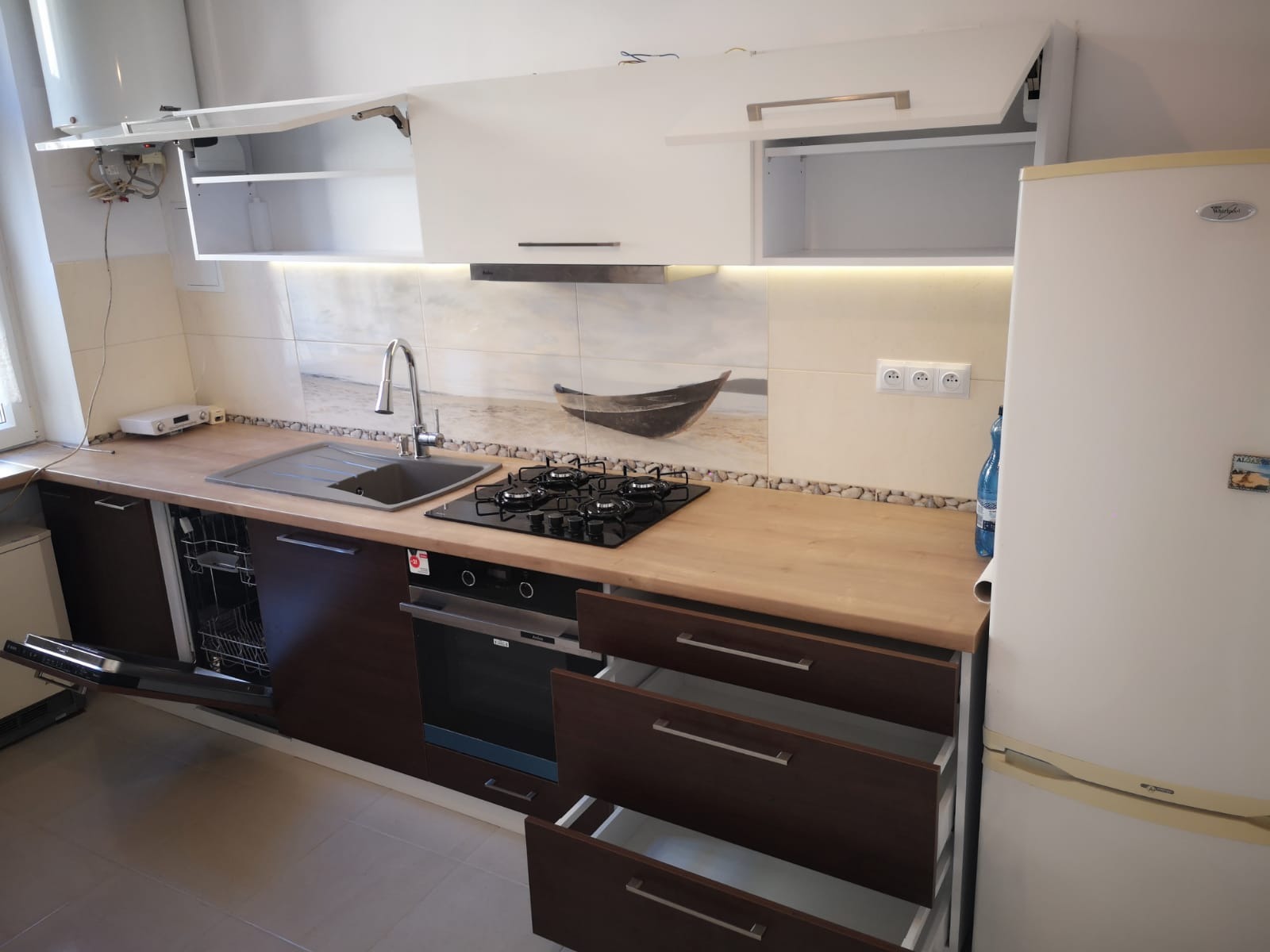 Nowoczesna kuchnia z drewnianymi blatami, ciemnobrązowymi szafkami z otwartymi szufladami, białymi szafkami wiszącymi z podświetleniem LED, zlewozmywakiem, płytą gazową i piekarnikiem, na ścianie...