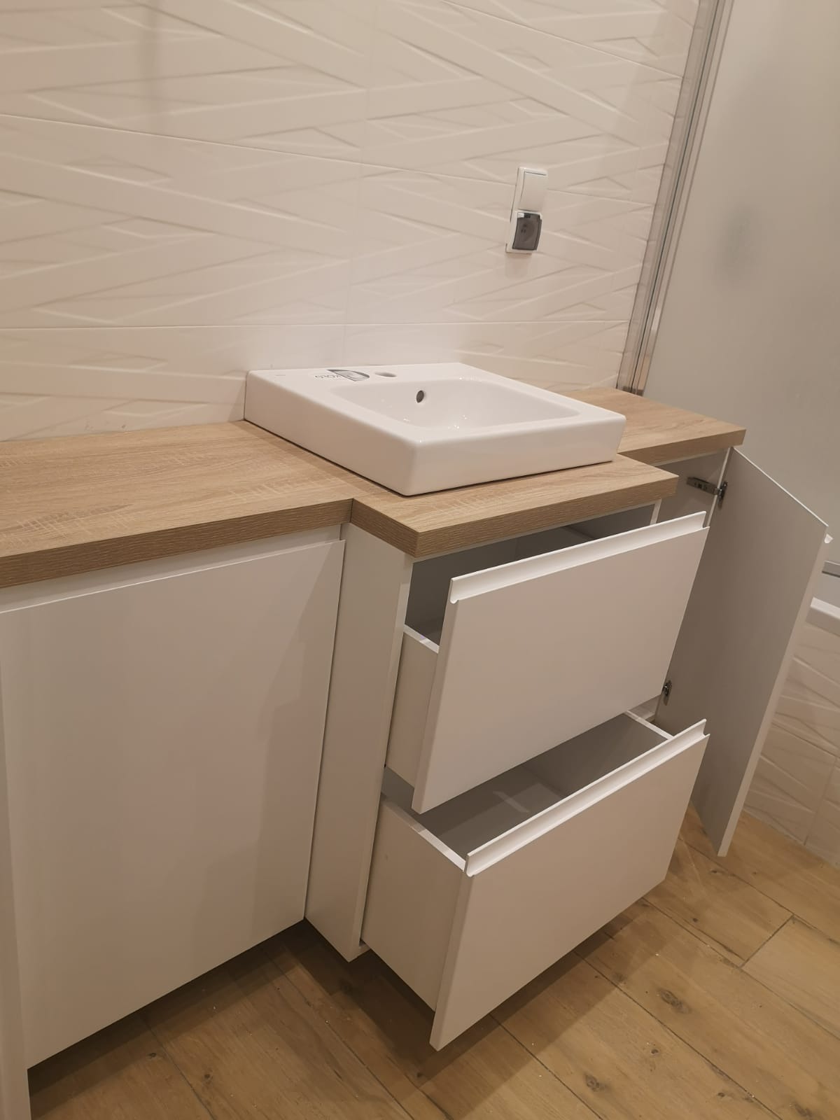 Biała umywalka nablatowa na szafce z blatem imitującym drewno, częściowo otwarte szuflady i drzwiczki, białe płytki na ścianie z delikatnym wzorem.