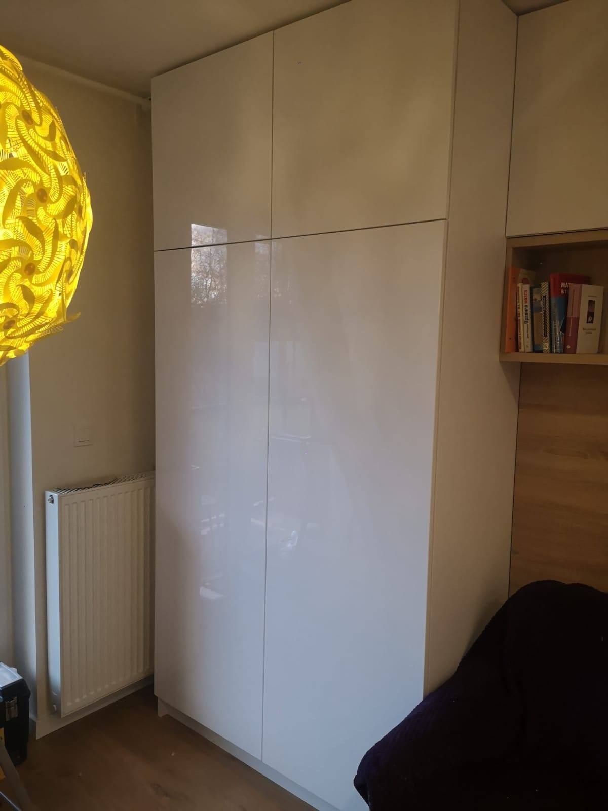Biała, wysoka szafa z połyskiem, z czterema drzwiami, zintegrowana z zabudową ścienną, widoczny grzejnik i żółta lampa z ażurowym abażurem.