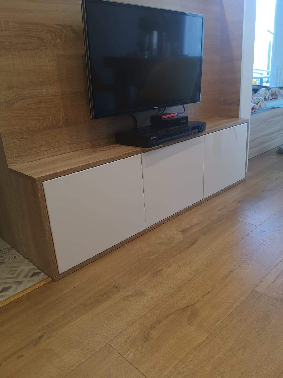 Telewizor zawieszony nad szafką RTV z białymi frontami i blatem imitującym drewno, na drewnianej podłodze.