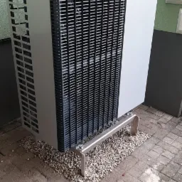 Szara pompa ciepła marki Viessmann na metalowych wspornikach, otoczona drobnymi kamieniami na tle zielonej ściany i szarego murku.