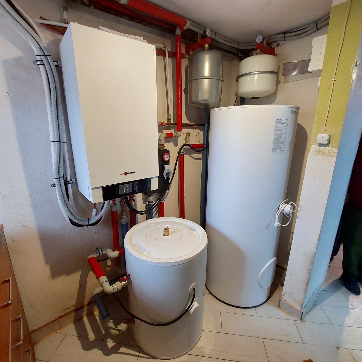 Kompaktowa kotłownia z białym piecem Viessmann, dwoma zbiornikami wyrównawczymi i dwoma zasobnikami ciepłej wody, rury w kolorze czerwonym i białym, instalacja w piwnicy.