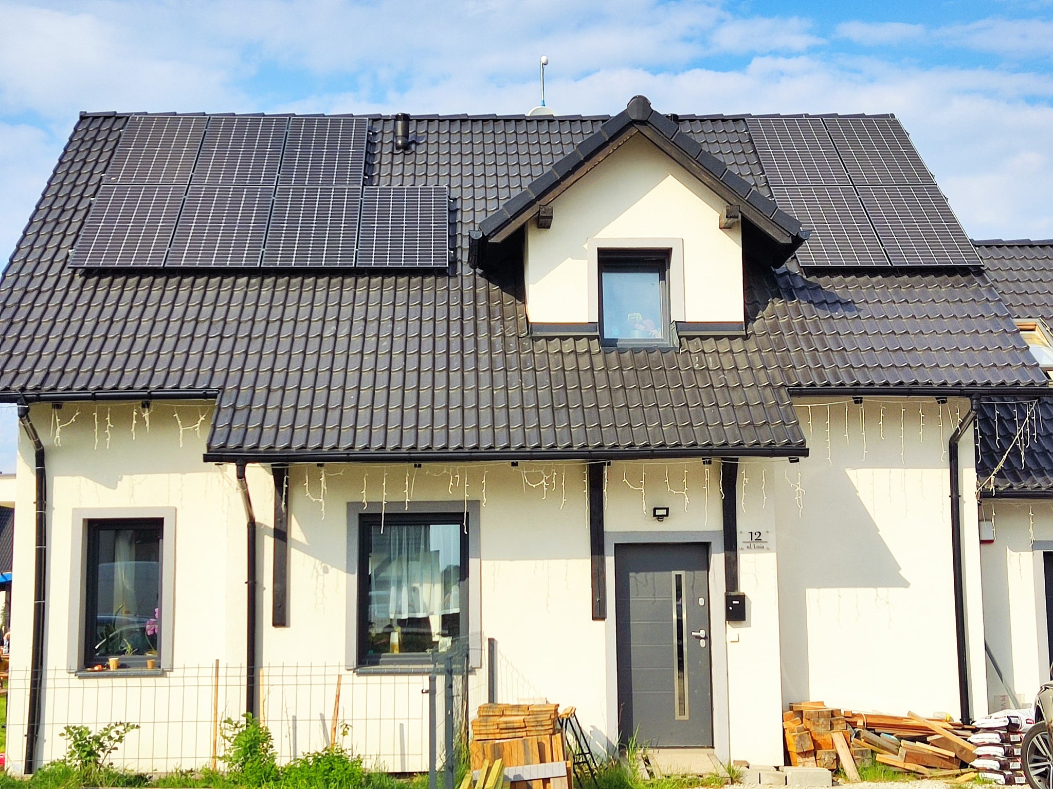 Biały dom jednorodzinny z ciemnoszarą dachówką i panelami słonecznymi na dachu, widok z zewnątrz w słoneczny dzień.