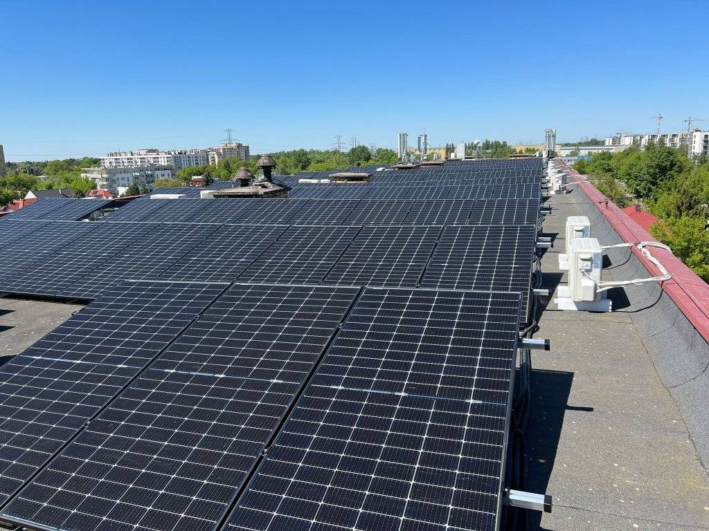 Rozległa instalacja paneli słonecznych na dachu budynku z widocznymi kominami wentylacyjnymi i jednostkami zewnętrznymi klimatyzacji, na tle miejskiego krajobrazu pod błękitnym niebem.