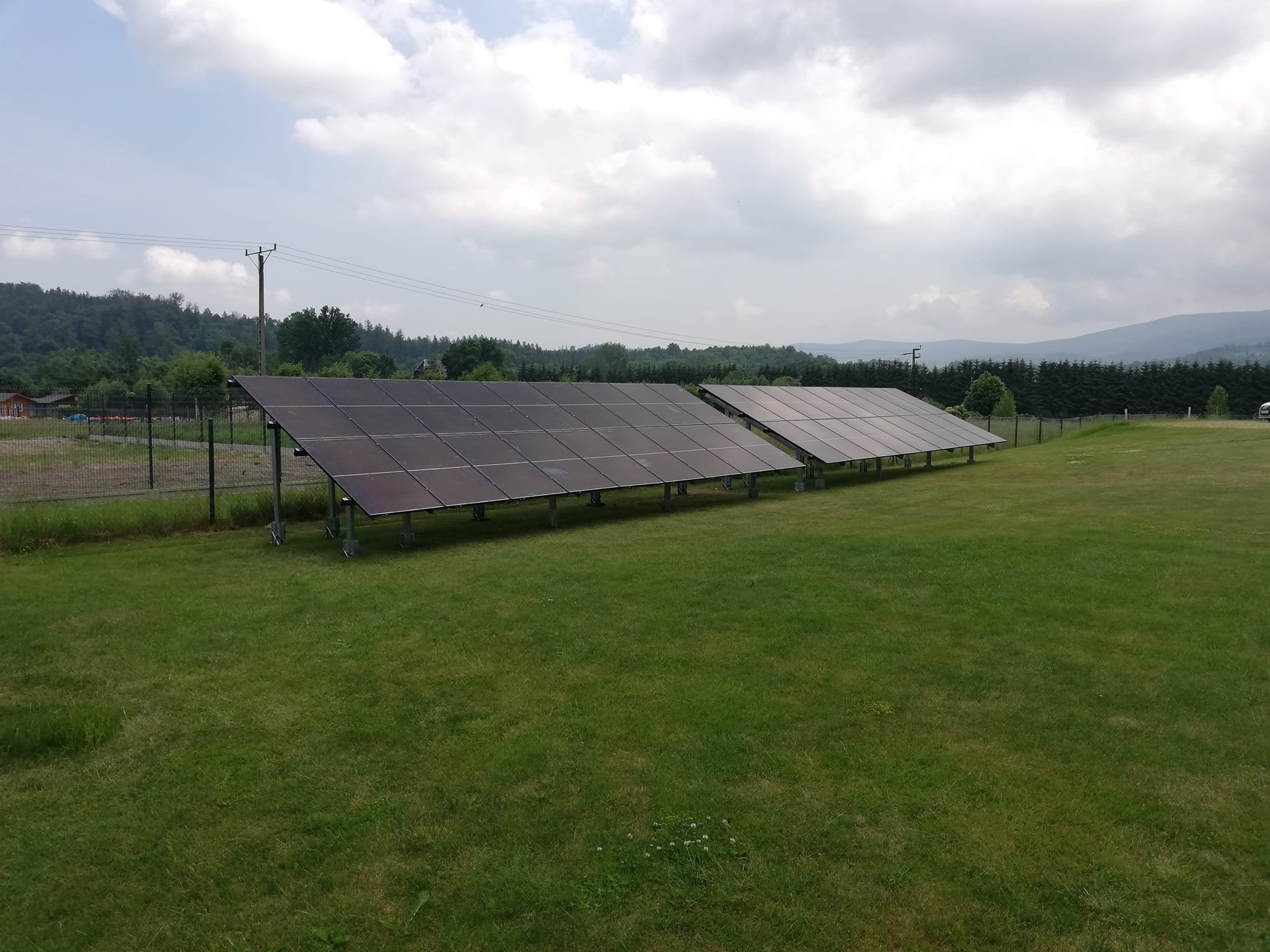 Dwa rzędy paneli słonecznych na metalowych konstrukcjach, zainstalowane na zielonej łące z widokiem na zalesione wzgórza i pochmurne niebo.