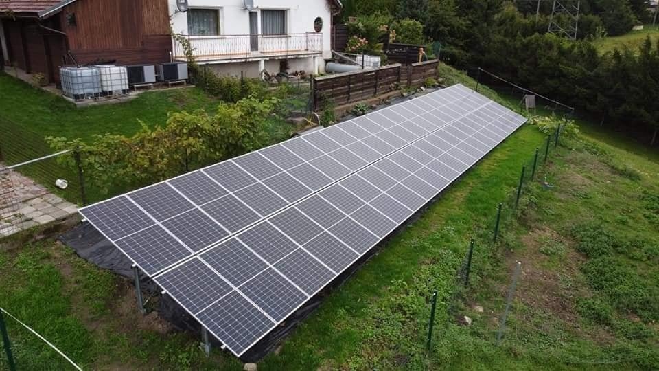 Długa instalacja paneli słonecznych na zboczu wzgórza, widoczna z góry, z domem w tle i metalowym ogrodzeniem po prawej stronie.