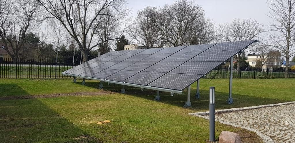 Instalacja paneli słonecznych na trawniku, widoczne metalowe podpory i otoczenie z drzewami i budynkami w tle.