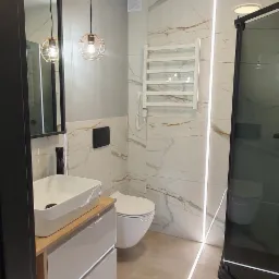 Nowoczesna łazienka z marmurowymi płytkami, podświetleniem LED w podłodze, wiszącą toaletą i umywalką nablatową na drewnianej szafce z szufladami, oraz lampami wiszącymi w metalowych kloszach.